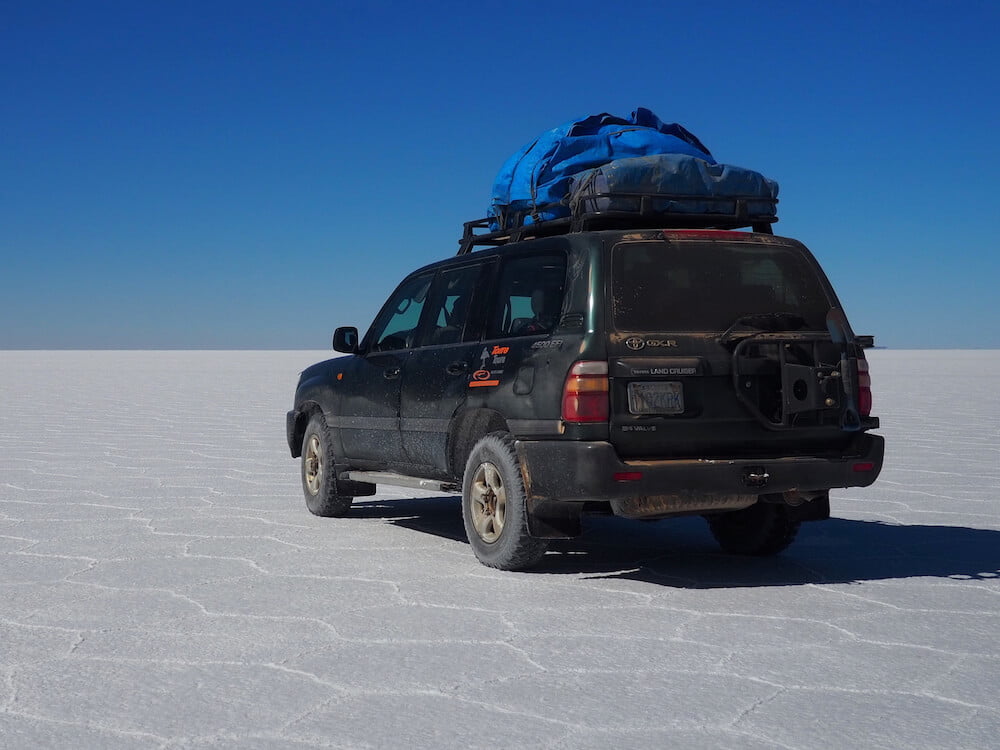 A jeep is parked on a flat white salt flat with blue sky above