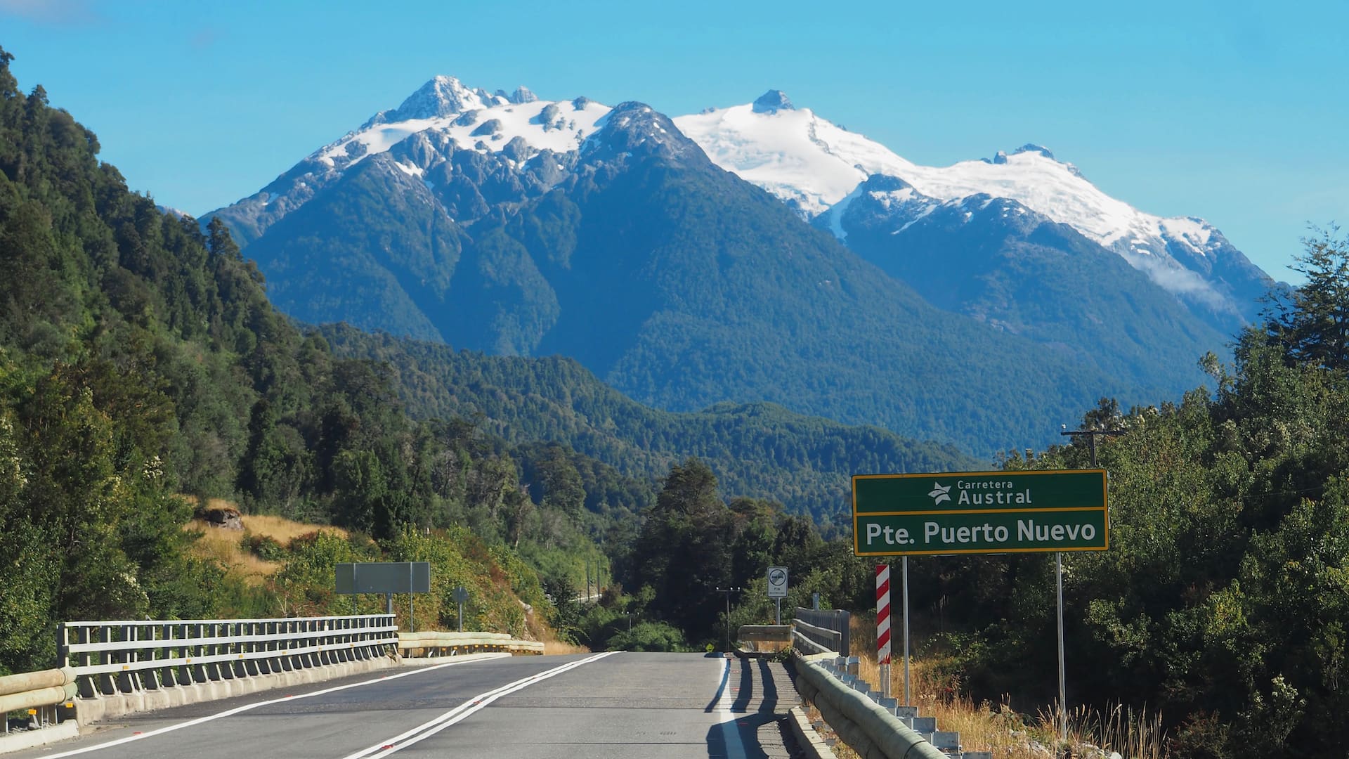 A green road sign stands beside a tarmac road with trees and a snow-capped mountain in the background