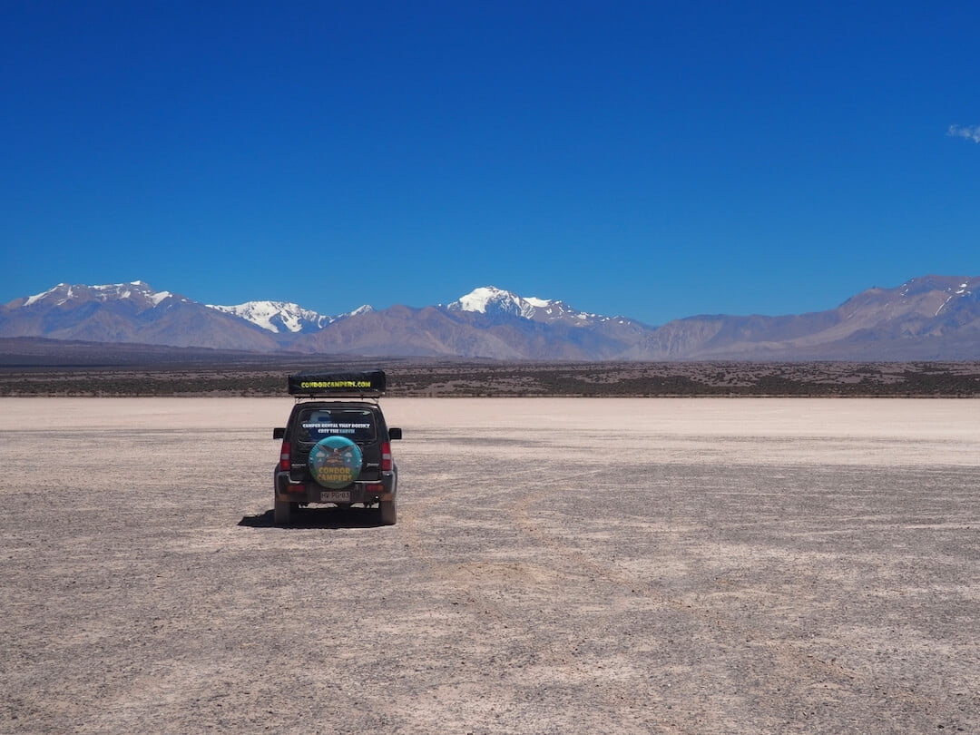 A jeep is parked on a dried mudflat with mountains in the distance and a blue sky