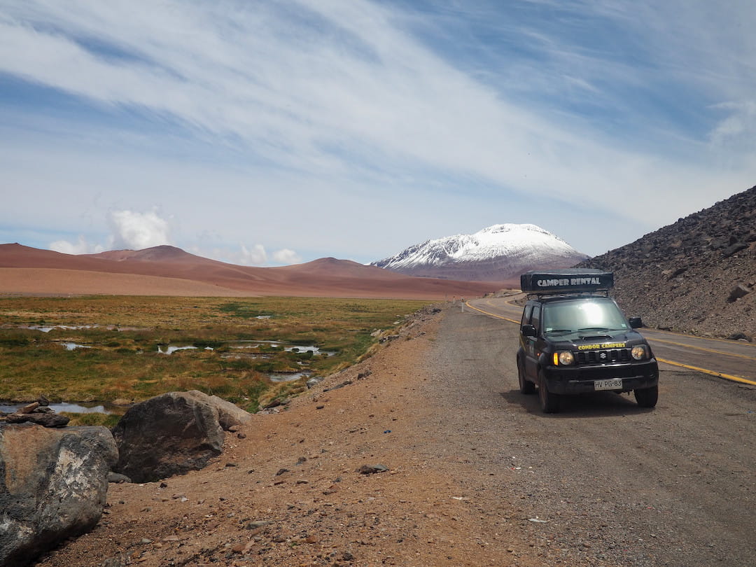 A jeep with a tent on its roof parked on a road with snow-capped mountain in the background