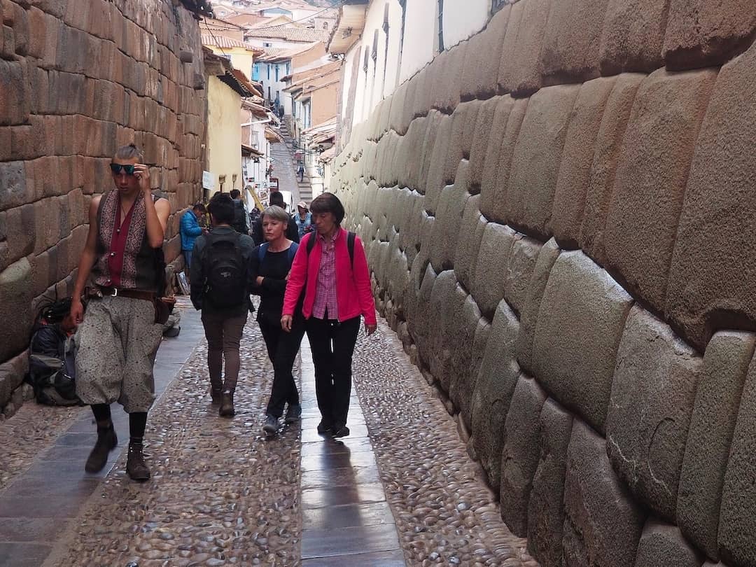 People walking in a narrow street with a large stone wall on the right