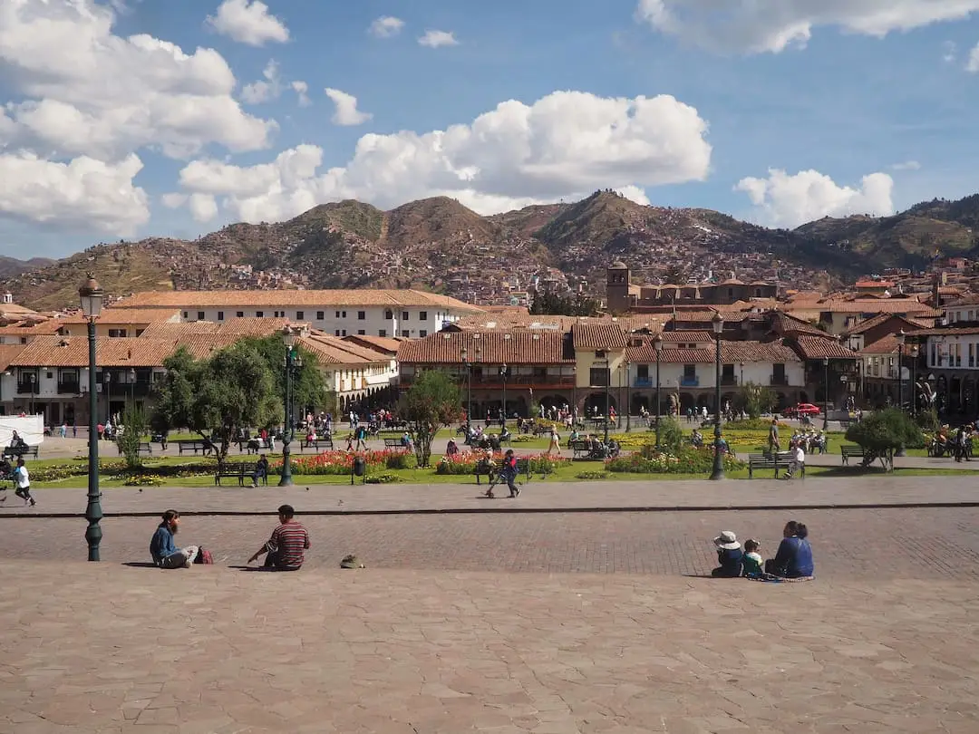 People sitting on stone steps in front of a square with buildings and mountains in the background