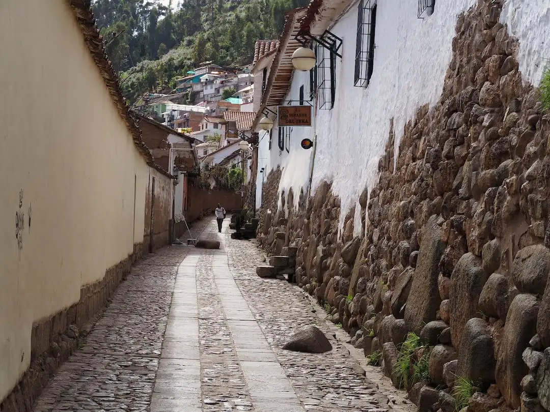 A narrow cobbled street with a long white-washed wall on the right half-covered in stone