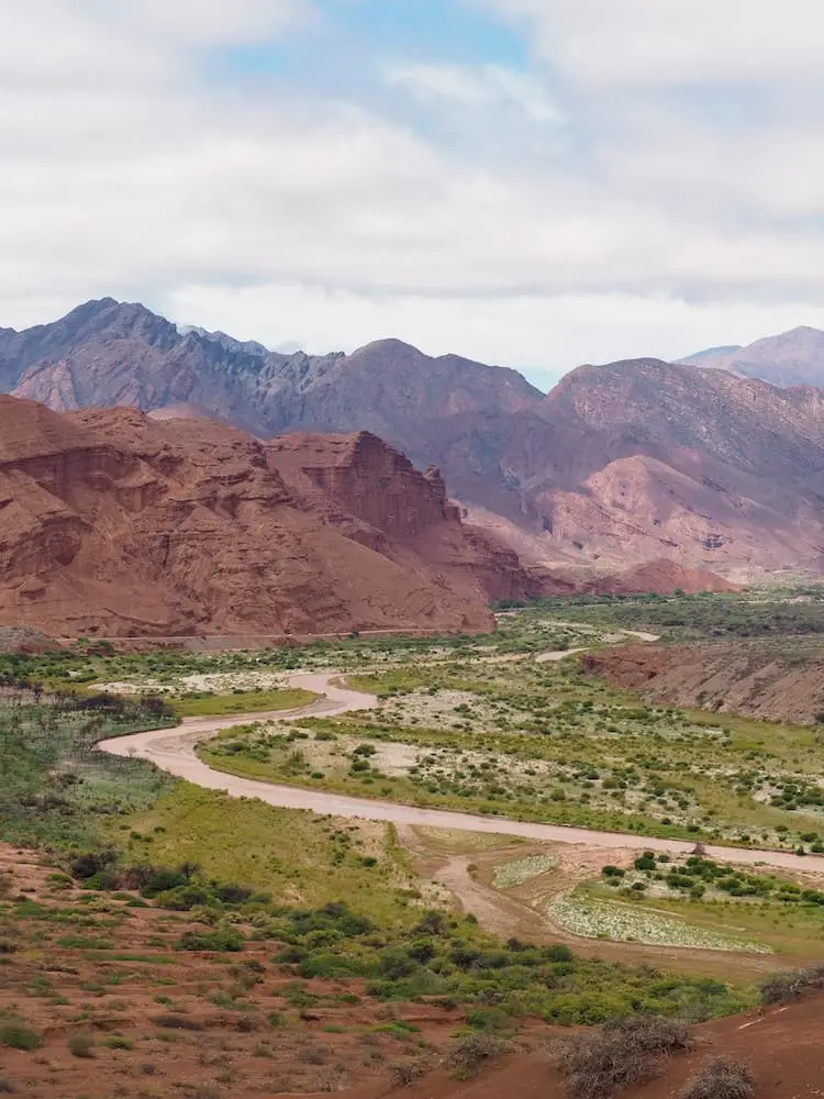 Winding road leading through a green valley floor with red-coloured mountains surrounding it