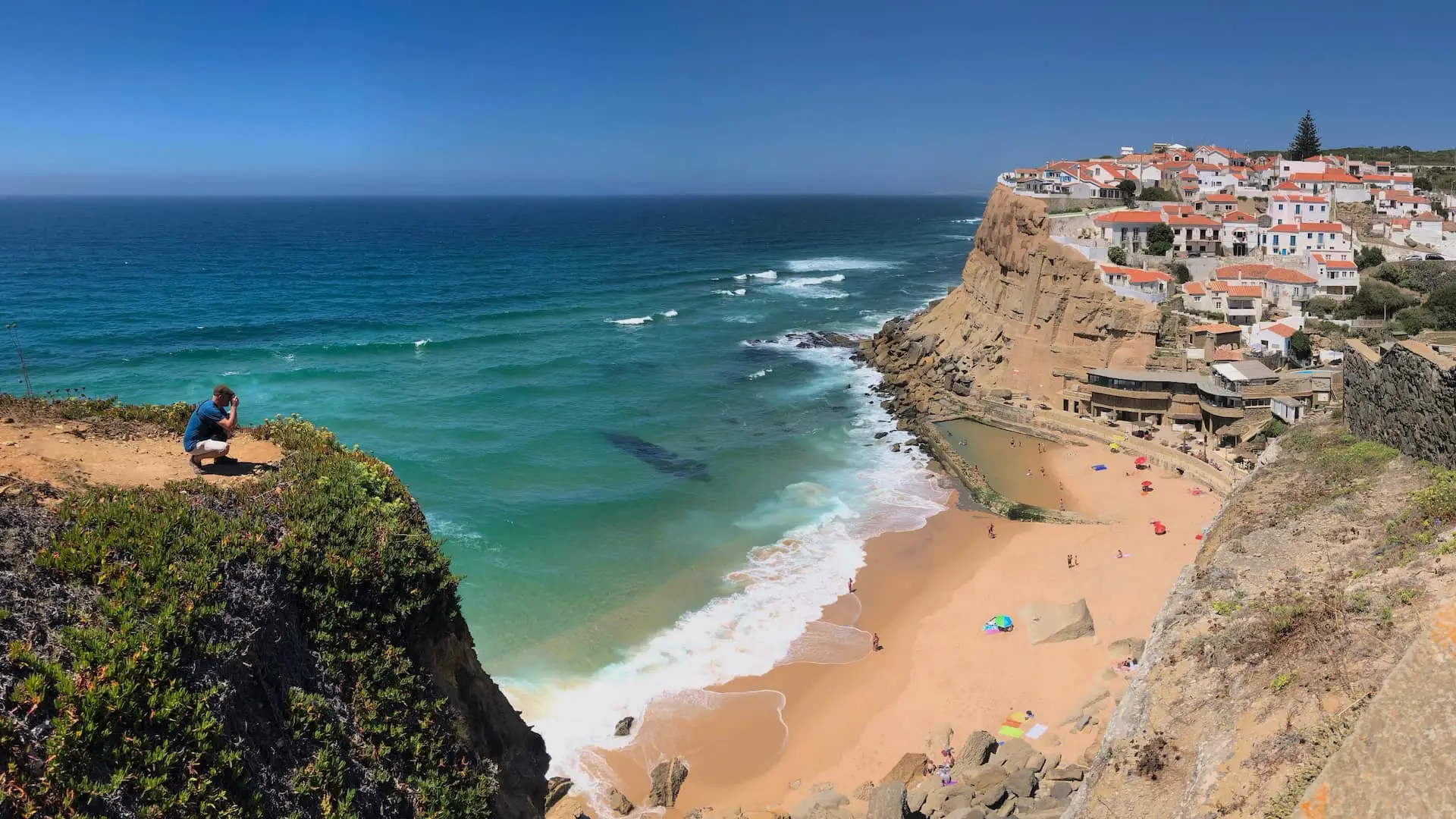 Ian crouches on a Rocky outcrop to the left to photograph, across a golden beach to a cliff-top village