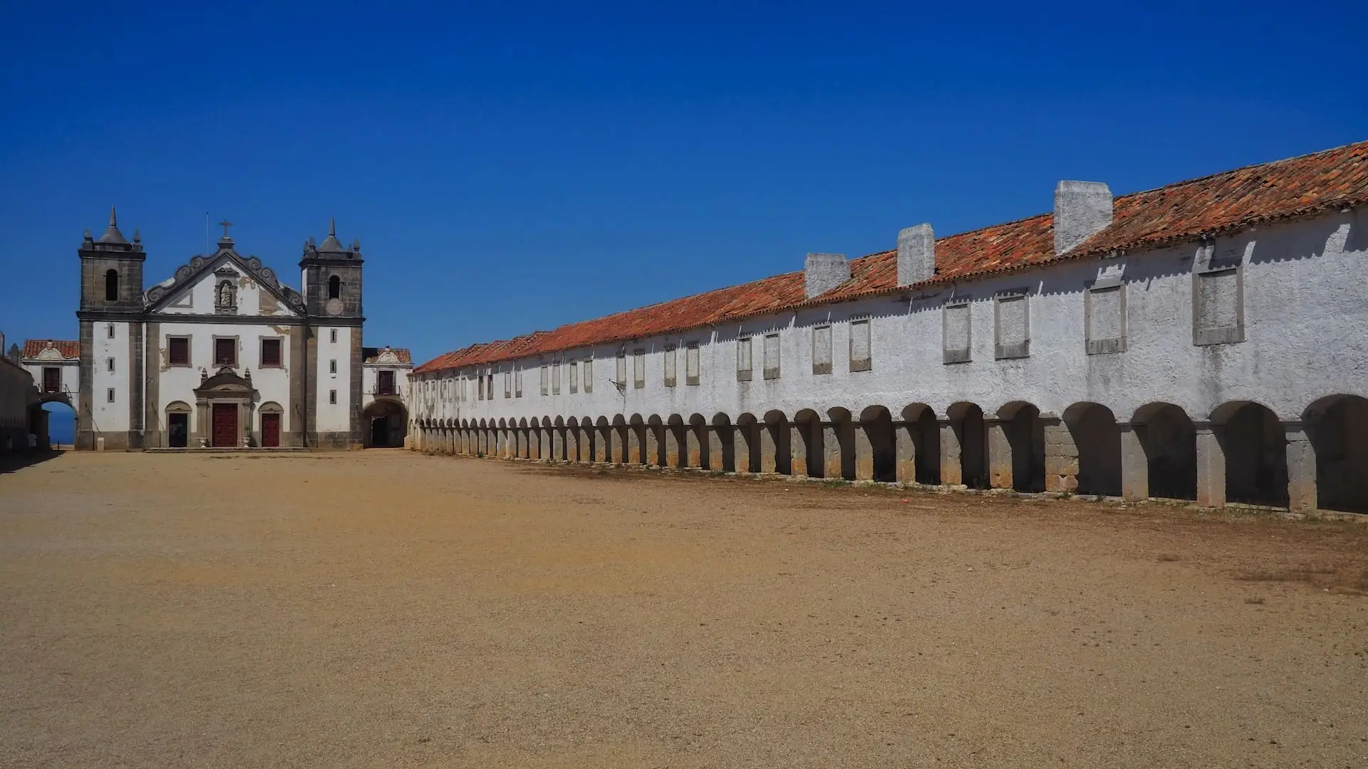 A long building with white wall, red roof and multiple arches leads to a church in the background