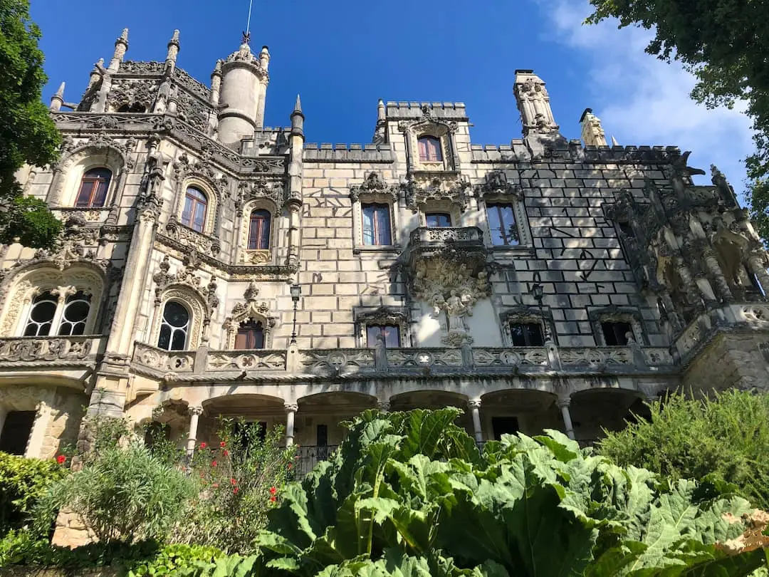 A white building with spires, arches and towers rises above green foliage