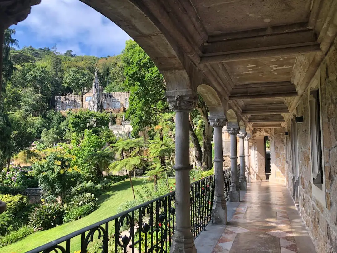 View from a terrace with stone pillars over to a building with a spire amidst lush gardens