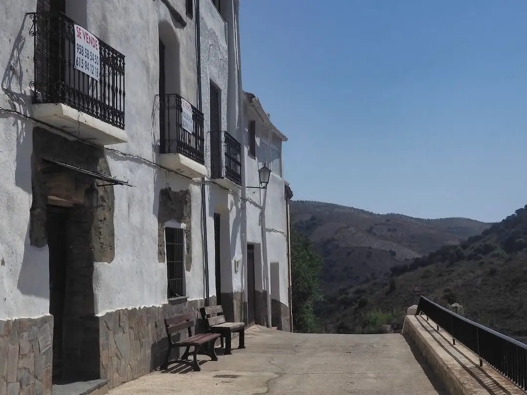 A row of terraced whitewashed buildings, two wooden benches out front, overlook a valley on the right