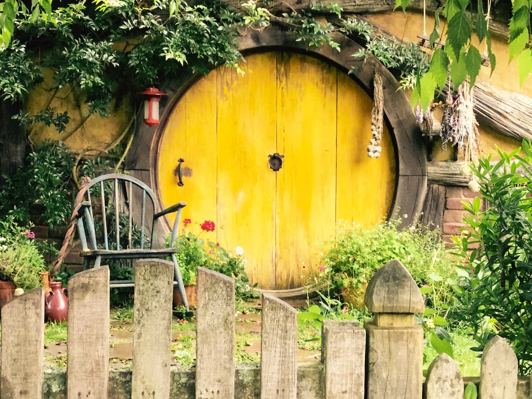 A circular yellow wooden door sits behind a garden fence and a wooden chair