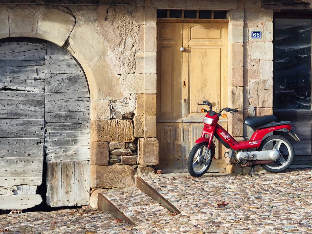 A red scooter rests against an old wall, next to a arched wooden doorway