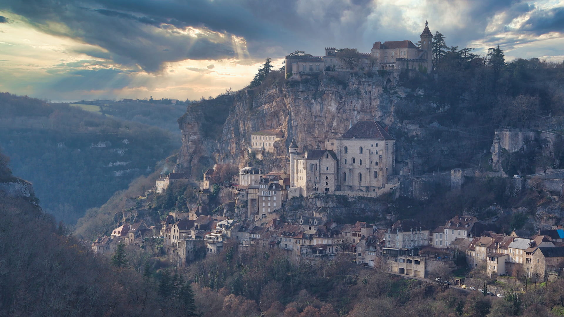 Stone houses cling to a hillside with a castle at the top, cloudy skies