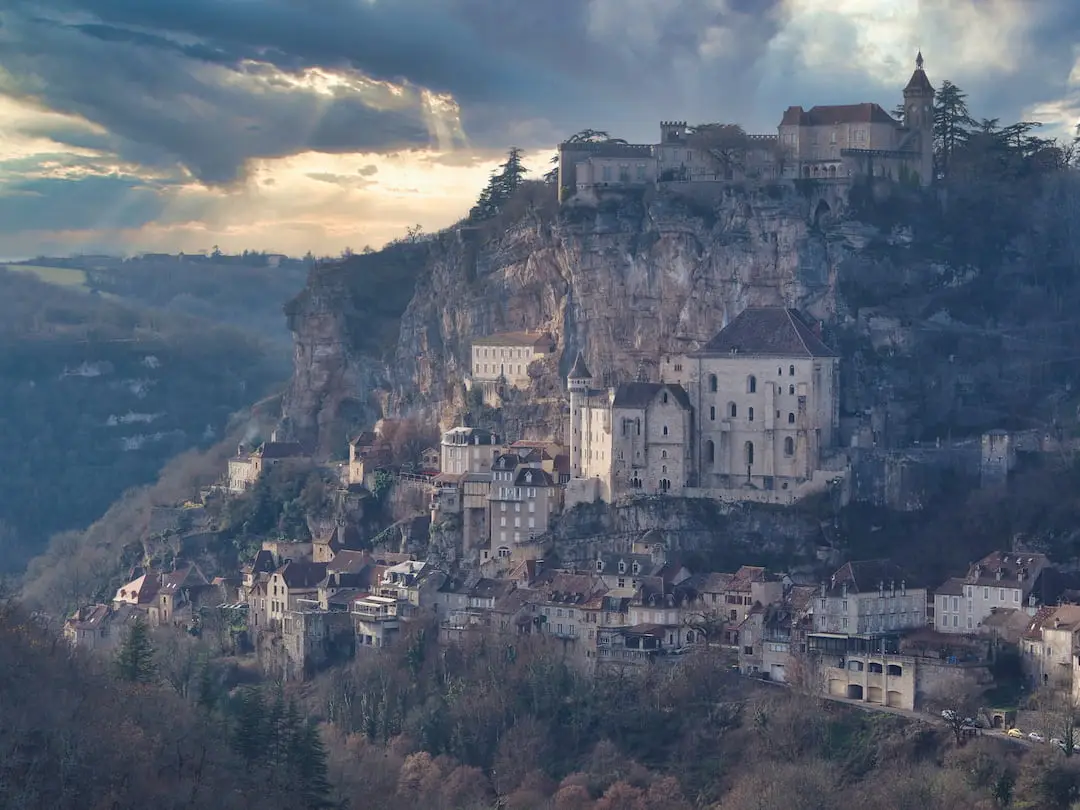 Stone houses cling to a hillside with a castle at the top, cloudy skies