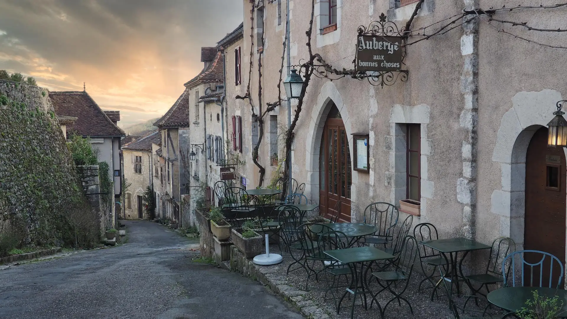 A street or stone houses runs downhill, right to left, to a sunset with green chairs in the foreground