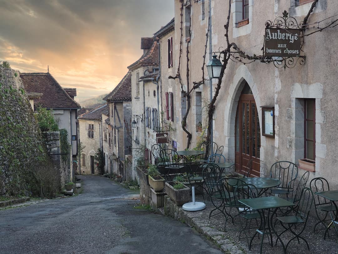 A street or stone houses runs downhill, right to left, to a sunset with green chairs in the foreground