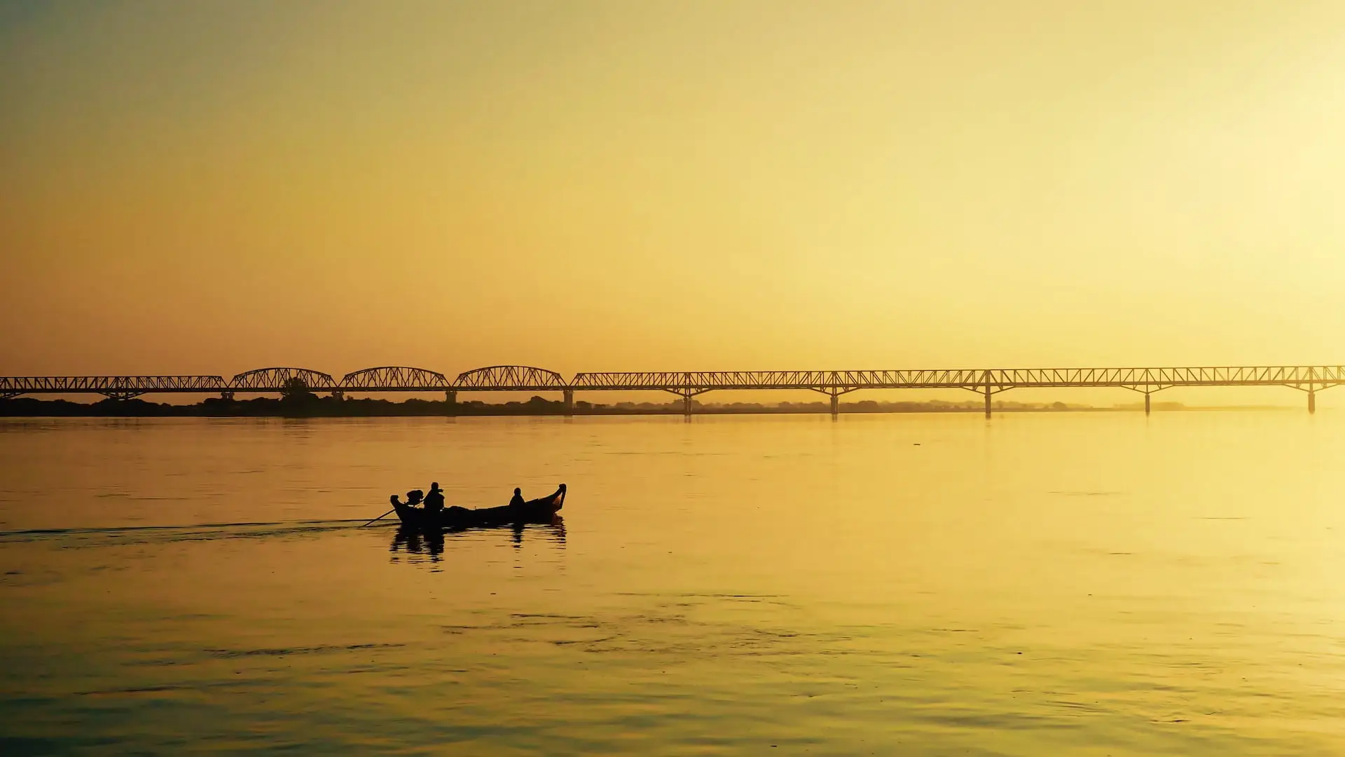 A small boat and a bridge are silhouetted against a yellow background of the sky and river