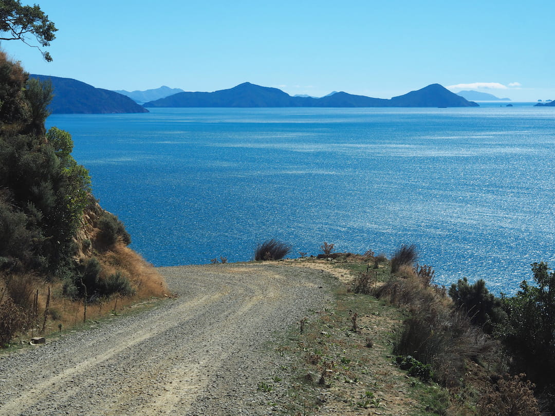 A gravel road turns to the left on a hillside overlooking the sea, with mountains in the distance