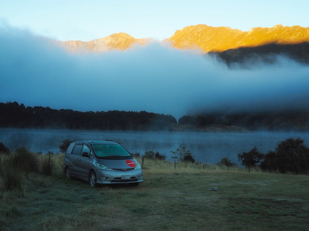 A car is parked on frosty ground beside a lake, with mist and mountains behind