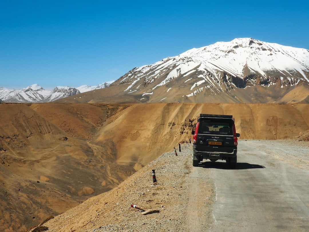 A car is parked on the side of a mountain road with a snow-capped peak in the background