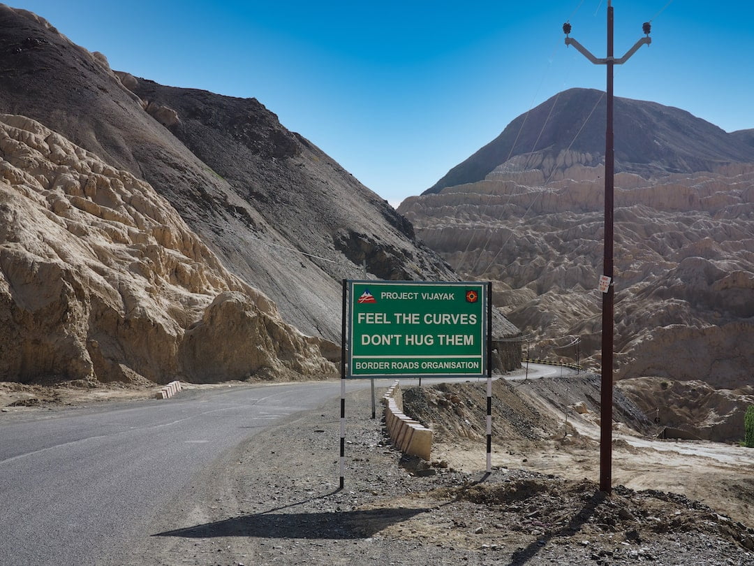 A green road sign with the words "Feel the curves, don't hug them" with mountains in the background