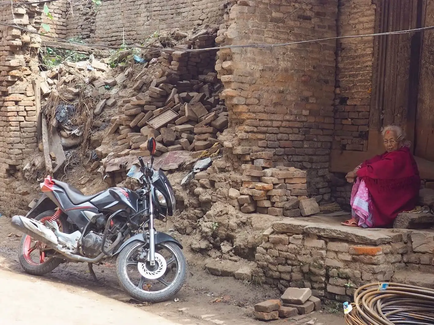 A woman in red clothing sits in a doorstep next to rubble and a motorcycle