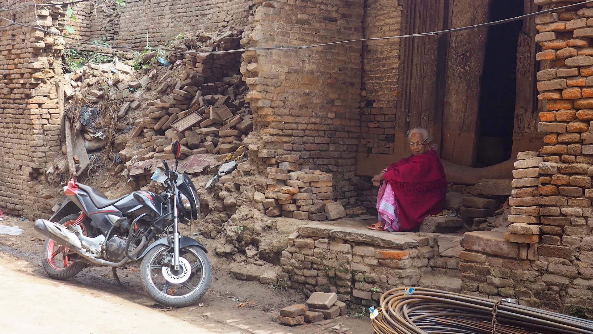 A woman in red clothing sits in a doorstep next to rubble and a motorcycle