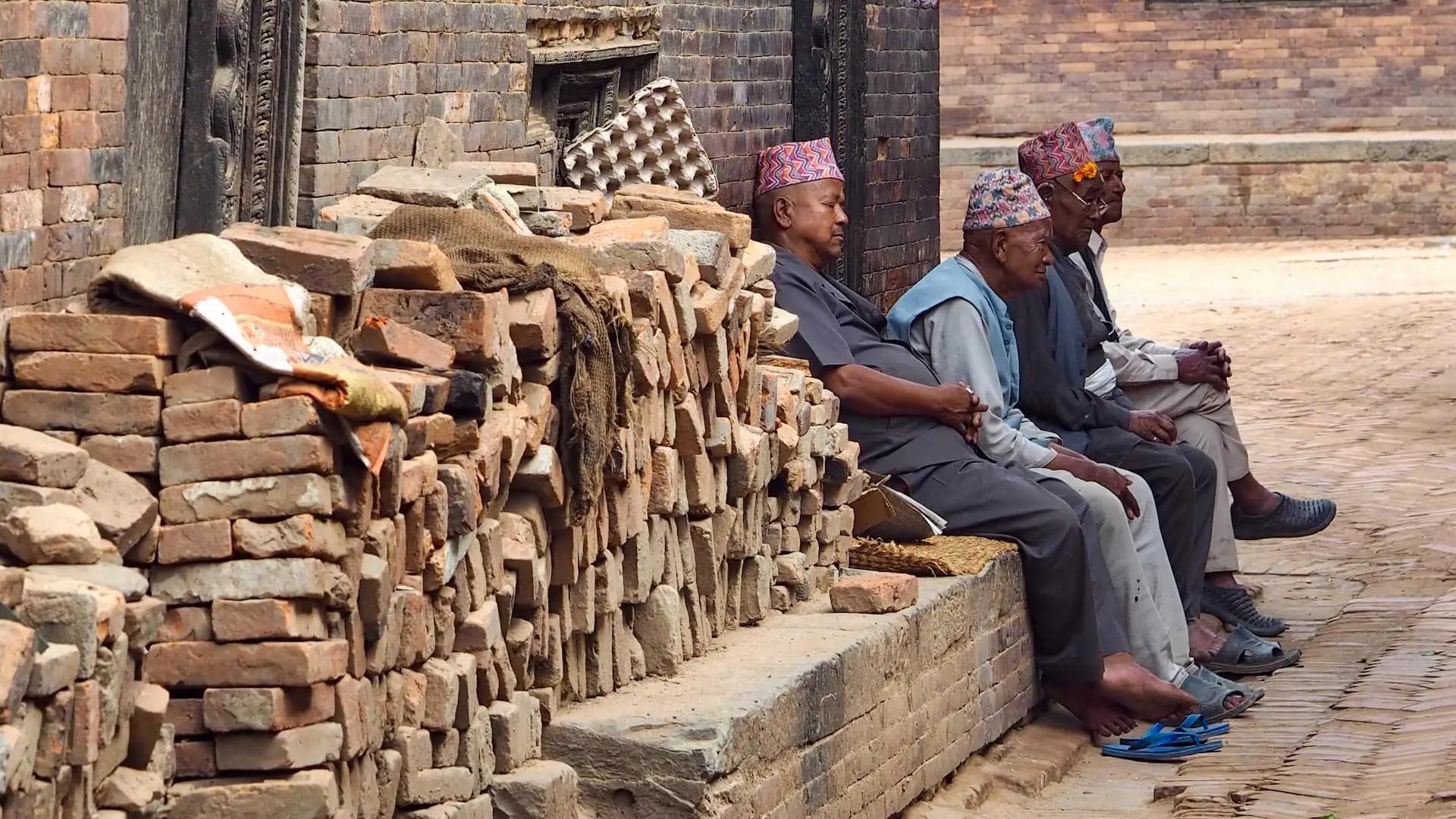 A pile of bricks in the foreground lead to four men sitting at the side of a street