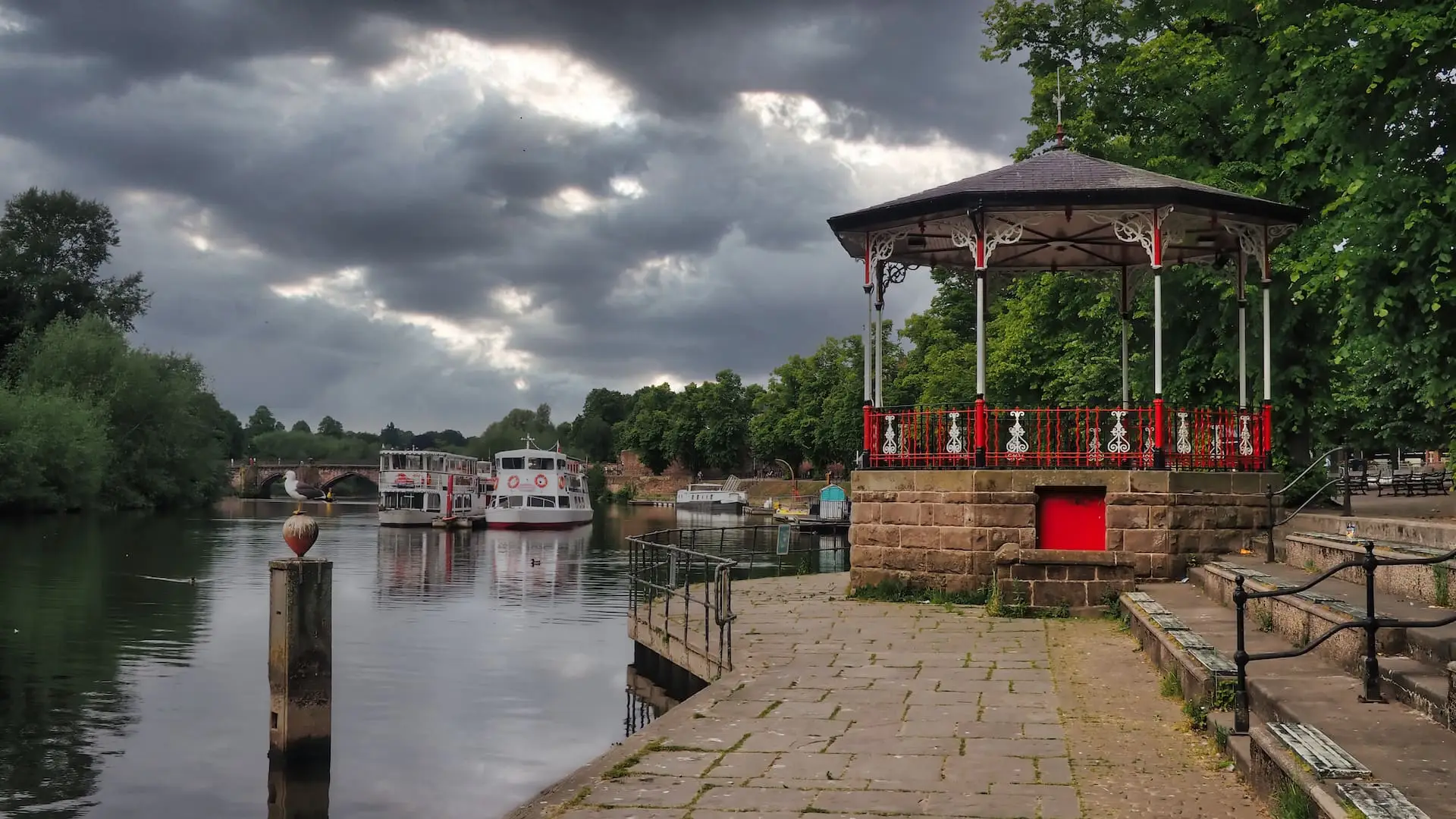 A red pagoda in the foreground stands next to a river with two boats in the background