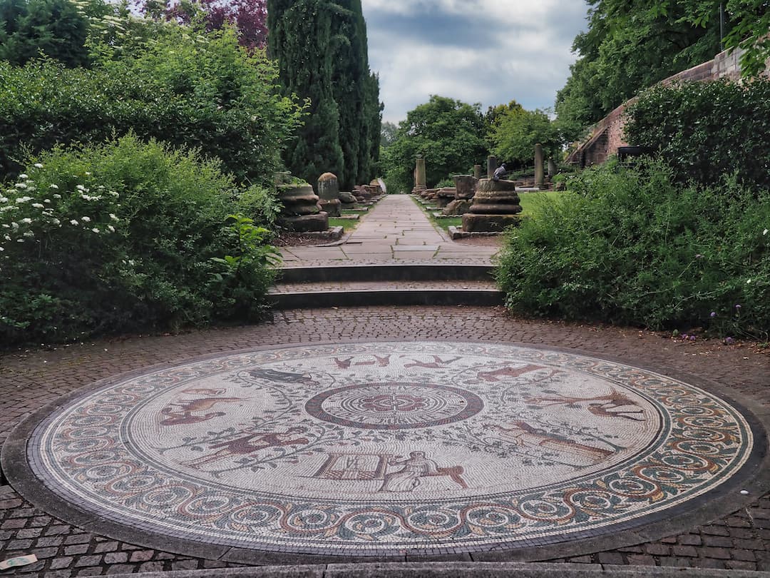 A circular mosaic in the foreground and a park with ancient stones in the background