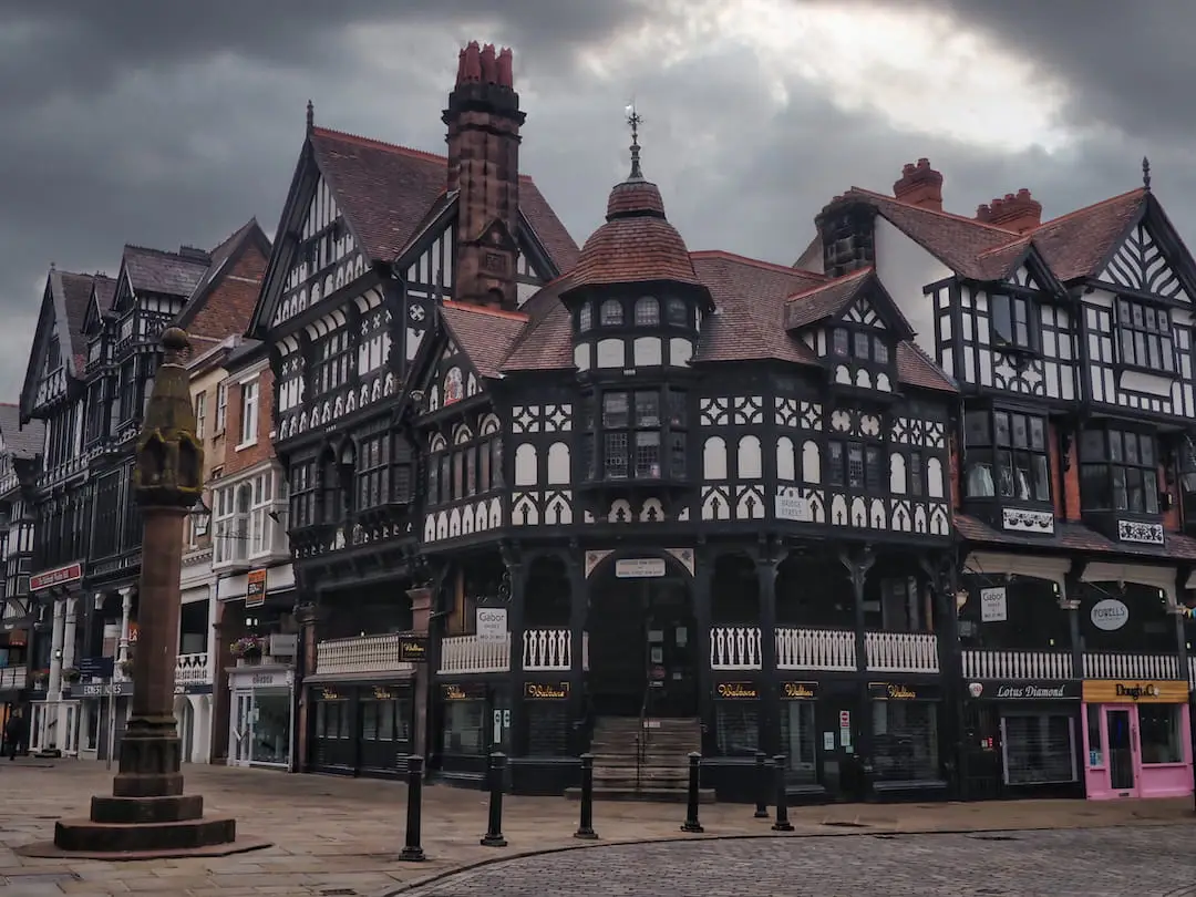 A sandstone cross on the left stands on a crossroads with black and white half-timbered buildings in the background
