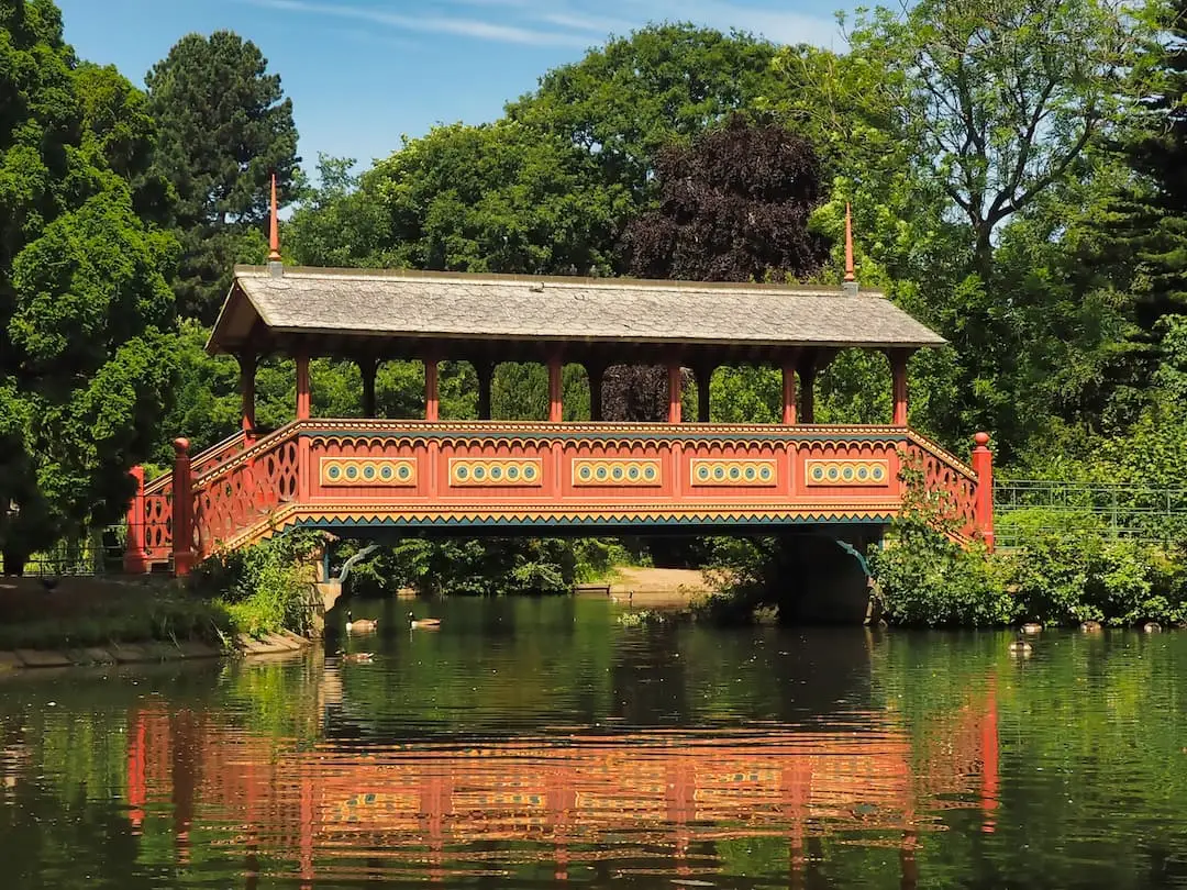 An red wooden bridge crosses a lake surrounded by ducks