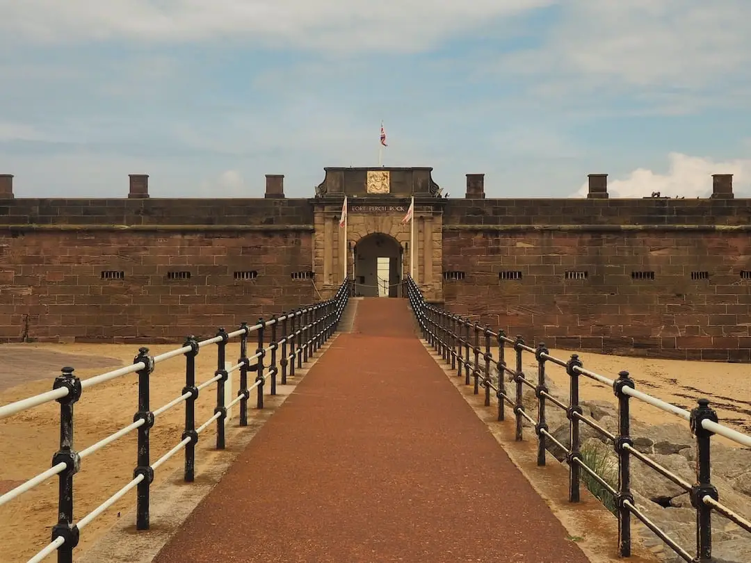 A red pathway border by railings leads to a gate at the head of a sandstone fort