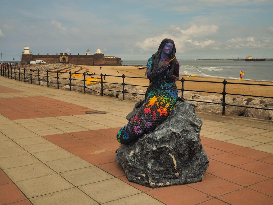 Sculpture of a mermaid on a promenade with Fort Perch Rock in the background