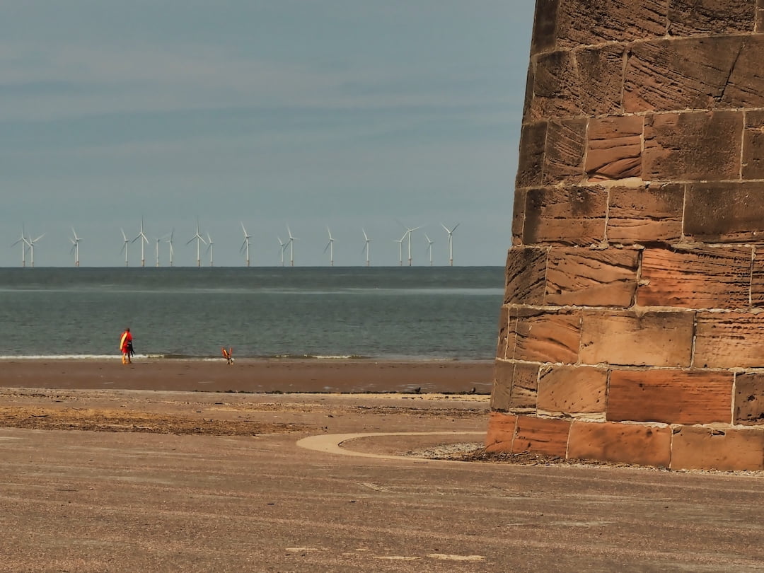 A red sandstone structure to the right, a beach, the sea and wind turbines to the left