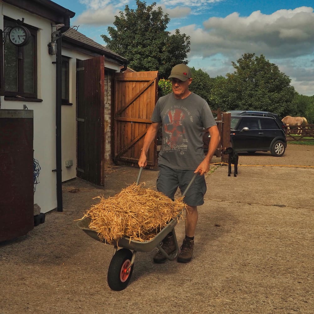 A man in a grey tee-shirt pushes a wheelbarrow filled with straw