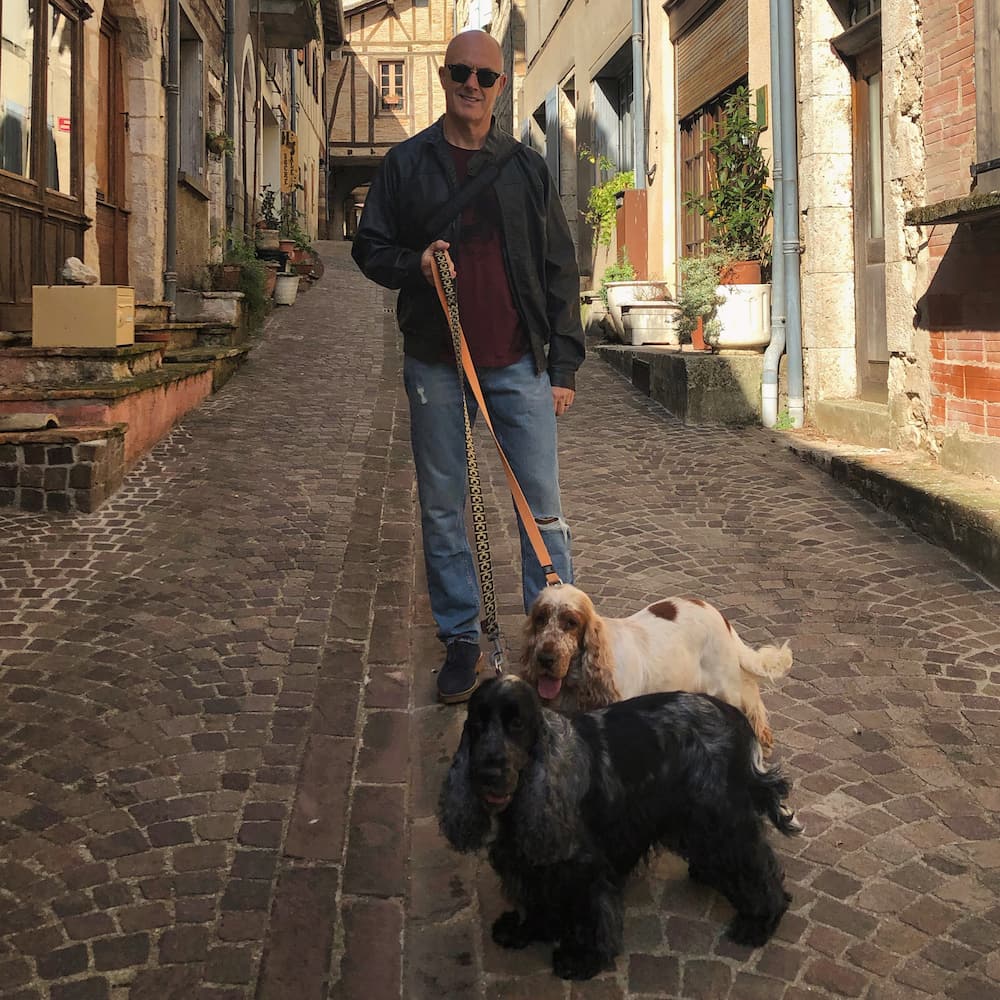 A man holds two dogs on a lead in a cobbled street surrounded by old buildings