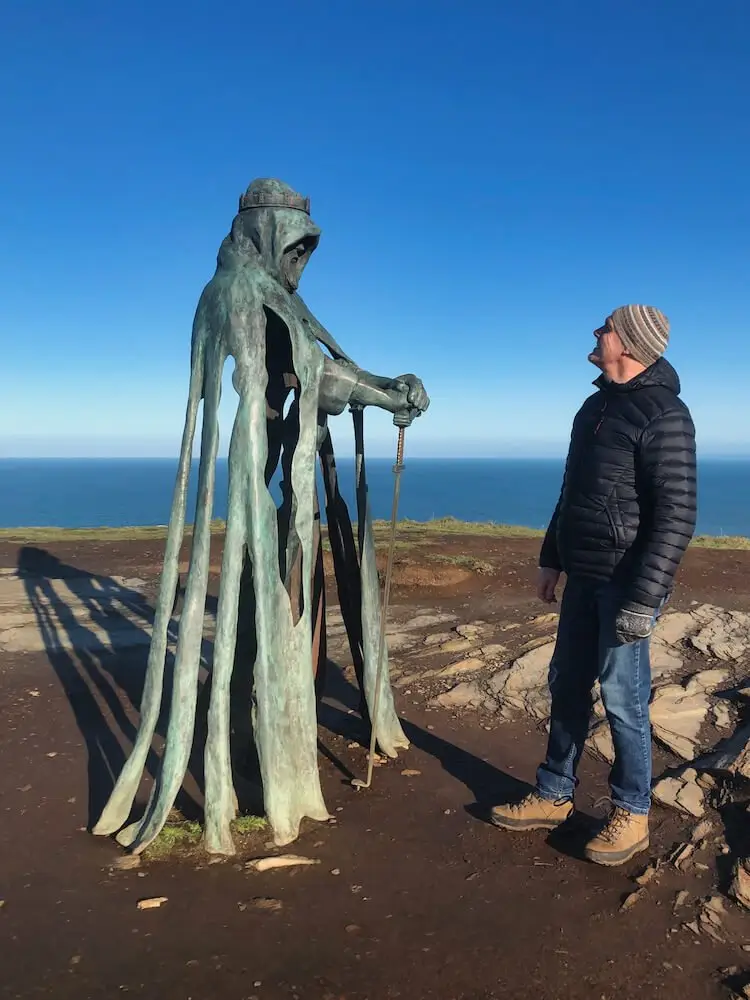 King Arthur Statue Cornwall. A statue of a man holding a sword on a clifftop with the sea in the background + a man staring up at it