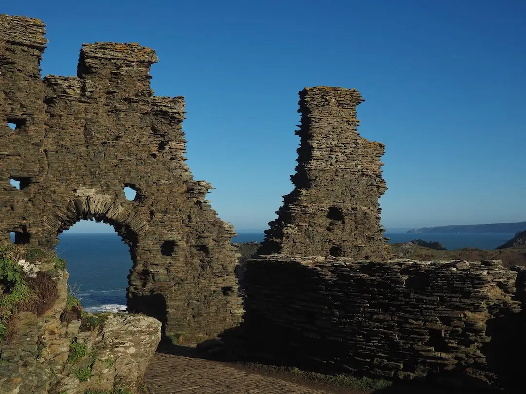 Tintagel Castle (1) A stone wall with an archway and small holes for windows crosses a pathway