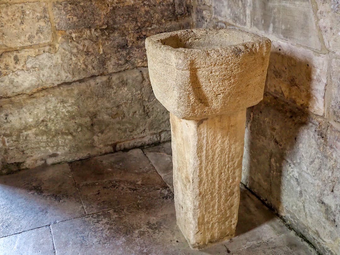 A stone structure, similar to a font, stands in the corner of a stone-built room