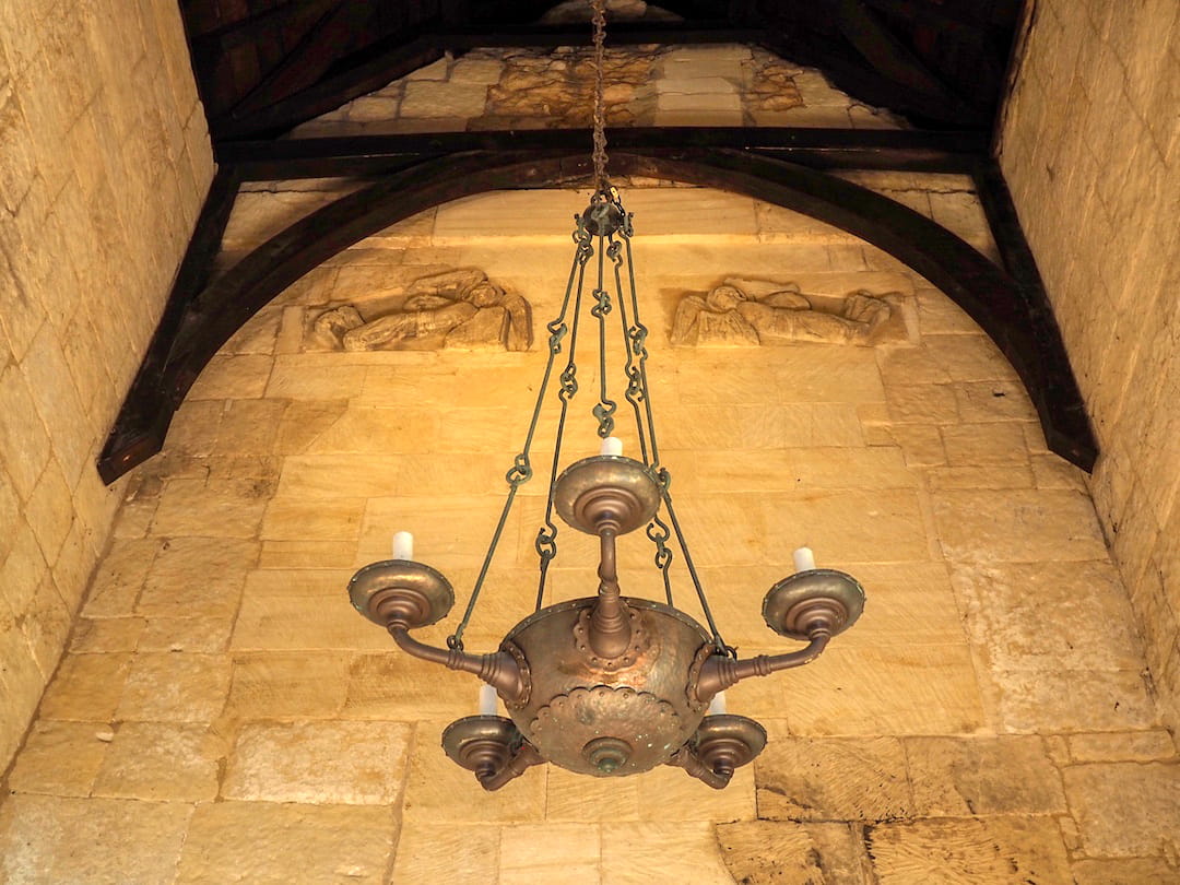 A chandelier hangs from a medieval ceiling in front of a stone interior wall