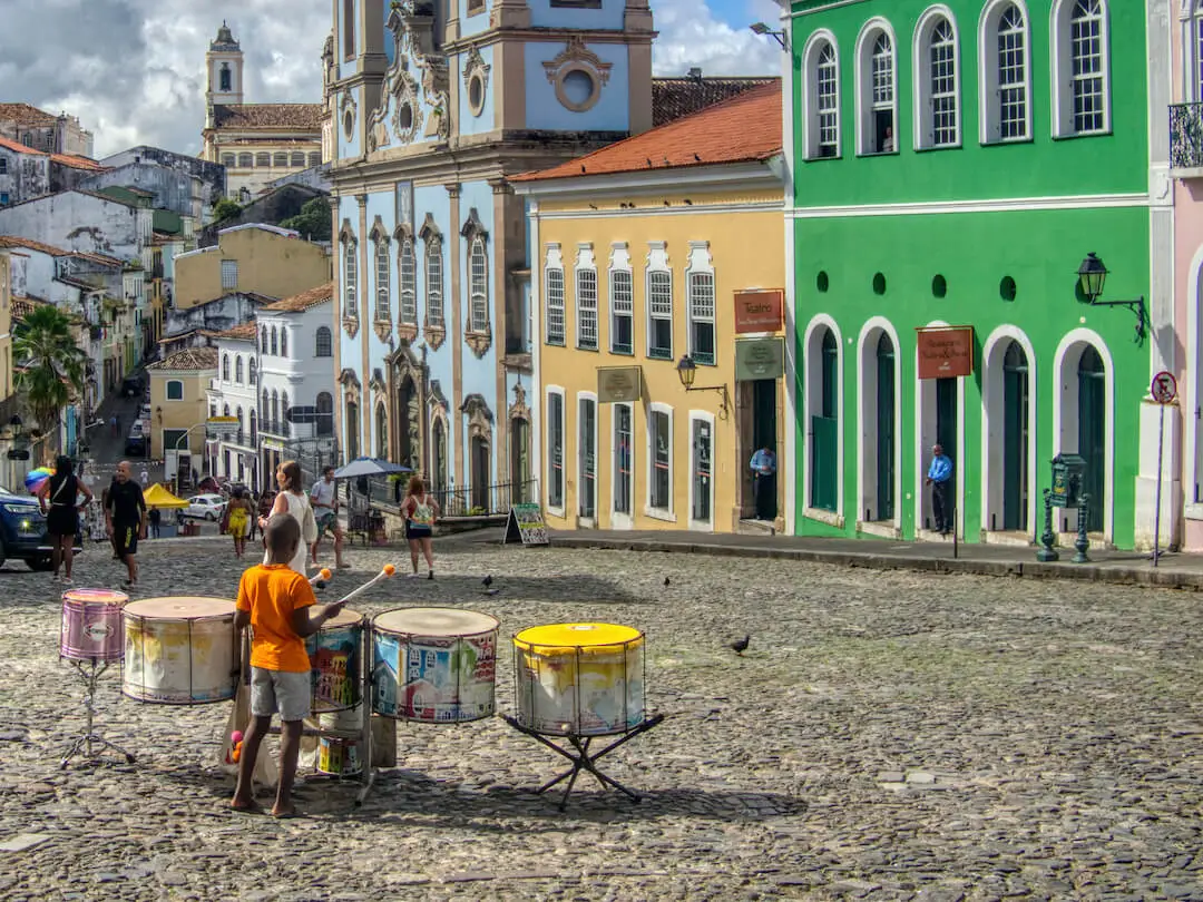 A small boy in an orange tee-shirt plays drums in a cobblestoned square