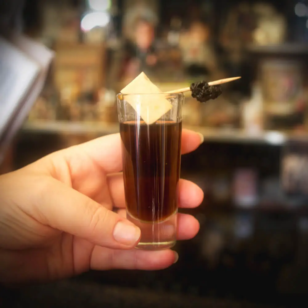 La Pasita Puebla A shot glass filled with an auburn-coloured drink, topped with a cube of white cheese on a stick
