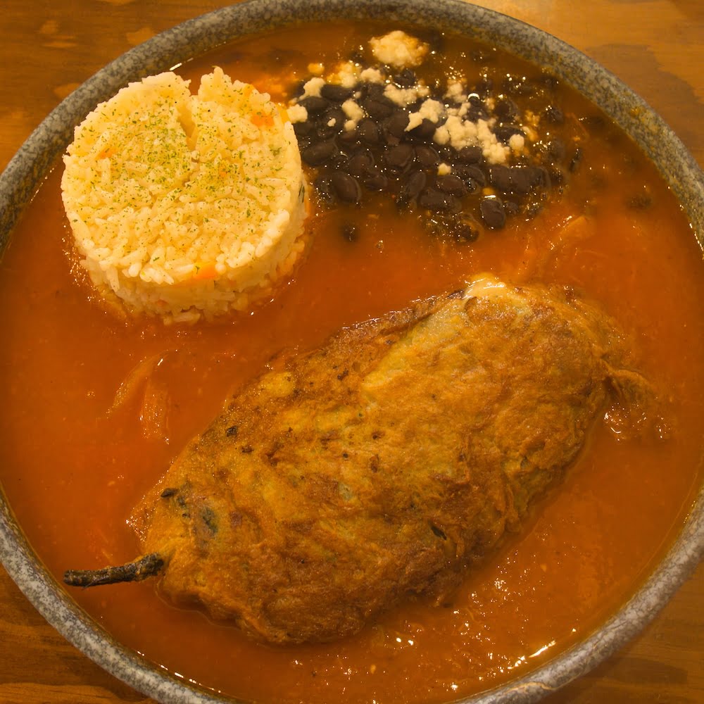 A bowl filled with red sauce, together with a stuffed chile, portion of rice and beans in 