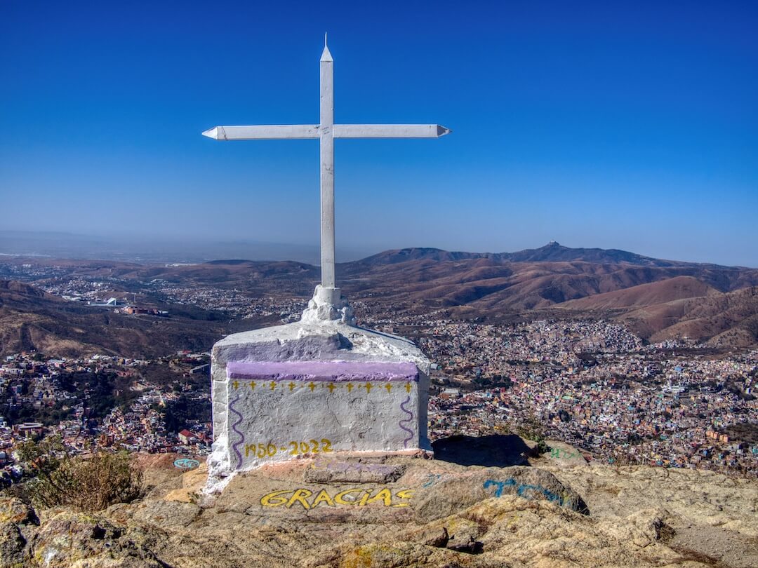 A white cross stands at the edge of a cliff overlooking a town