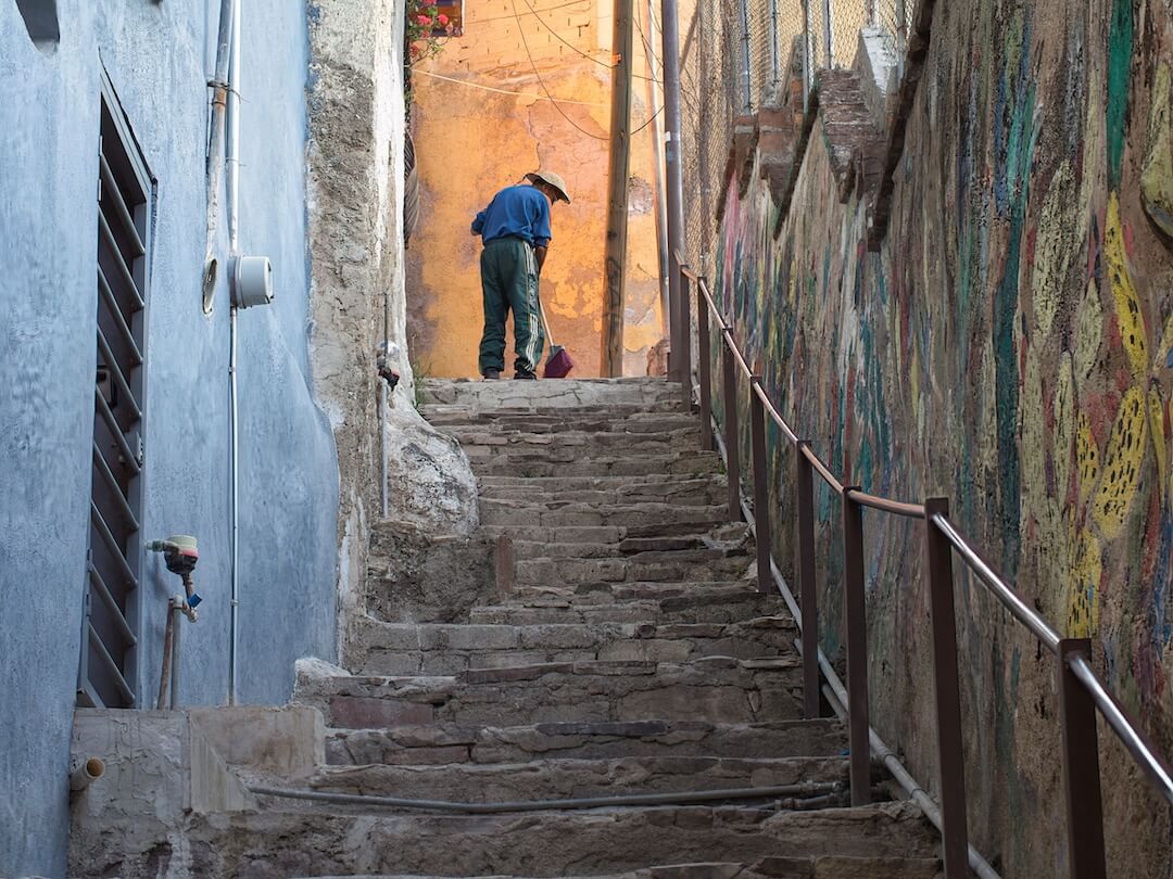 A man sweeps the pavement in a narrow alley at the top of a set of stone steps