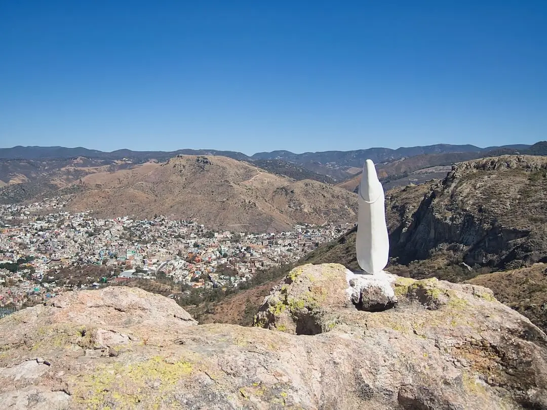  A slim, white statue looks out over a valley from a cliff edge