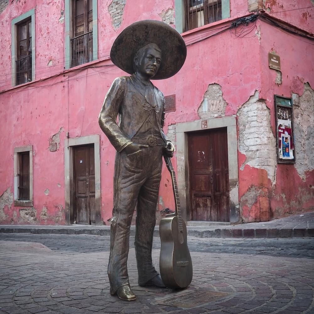 A bronze statue of a man with a guitar in front of a pink building