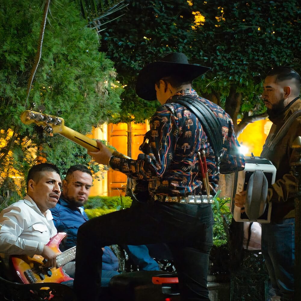 A man with a guitar stands facing another man on a bench