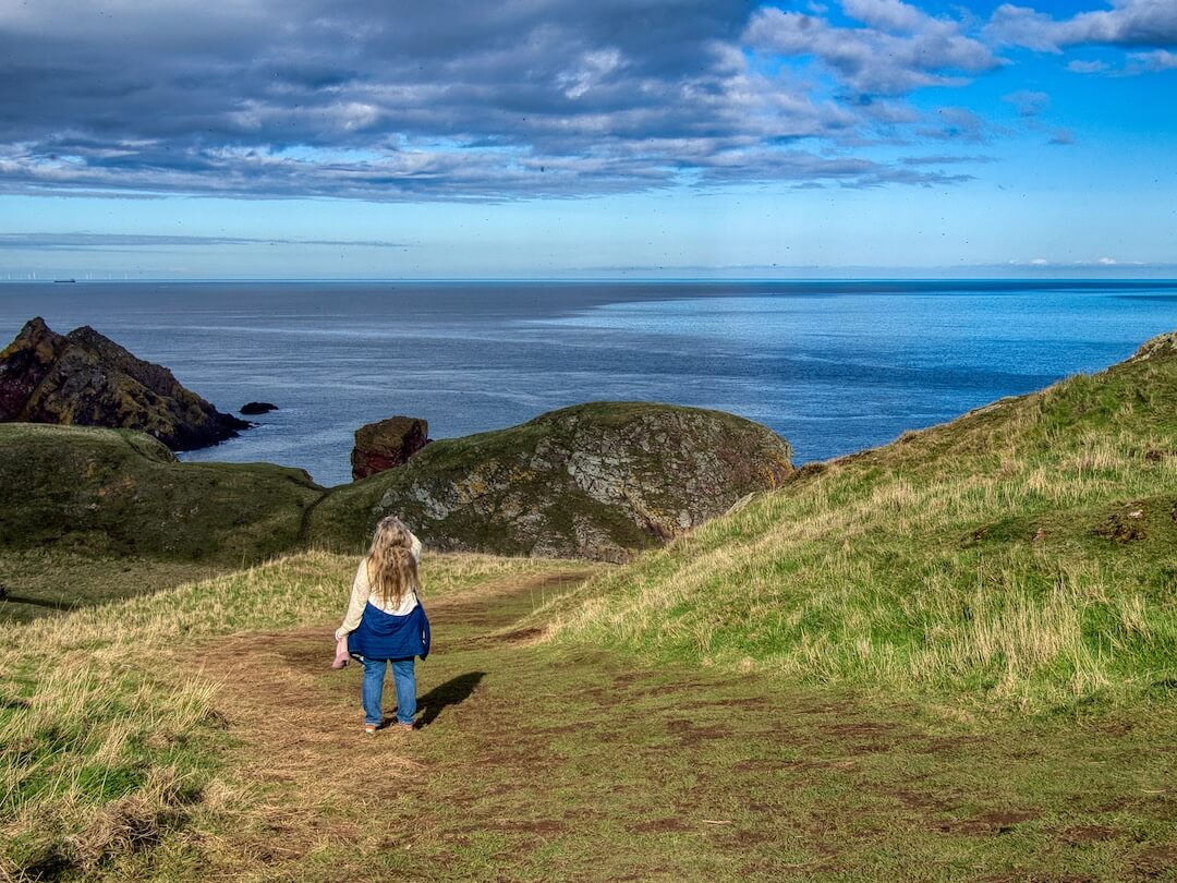 Nicky stands (looking away) on a footpath beside coastal cliffs 