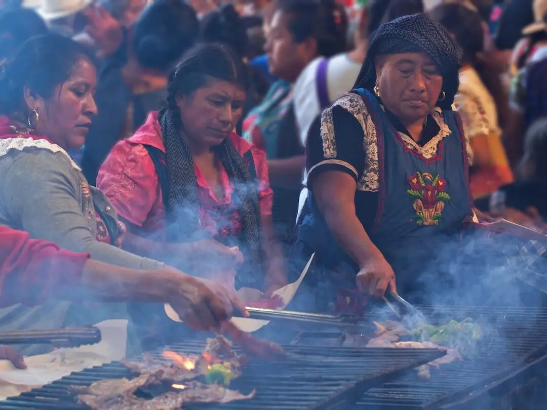 Three women in traditional dress cook meat and vegetables on a BBQ